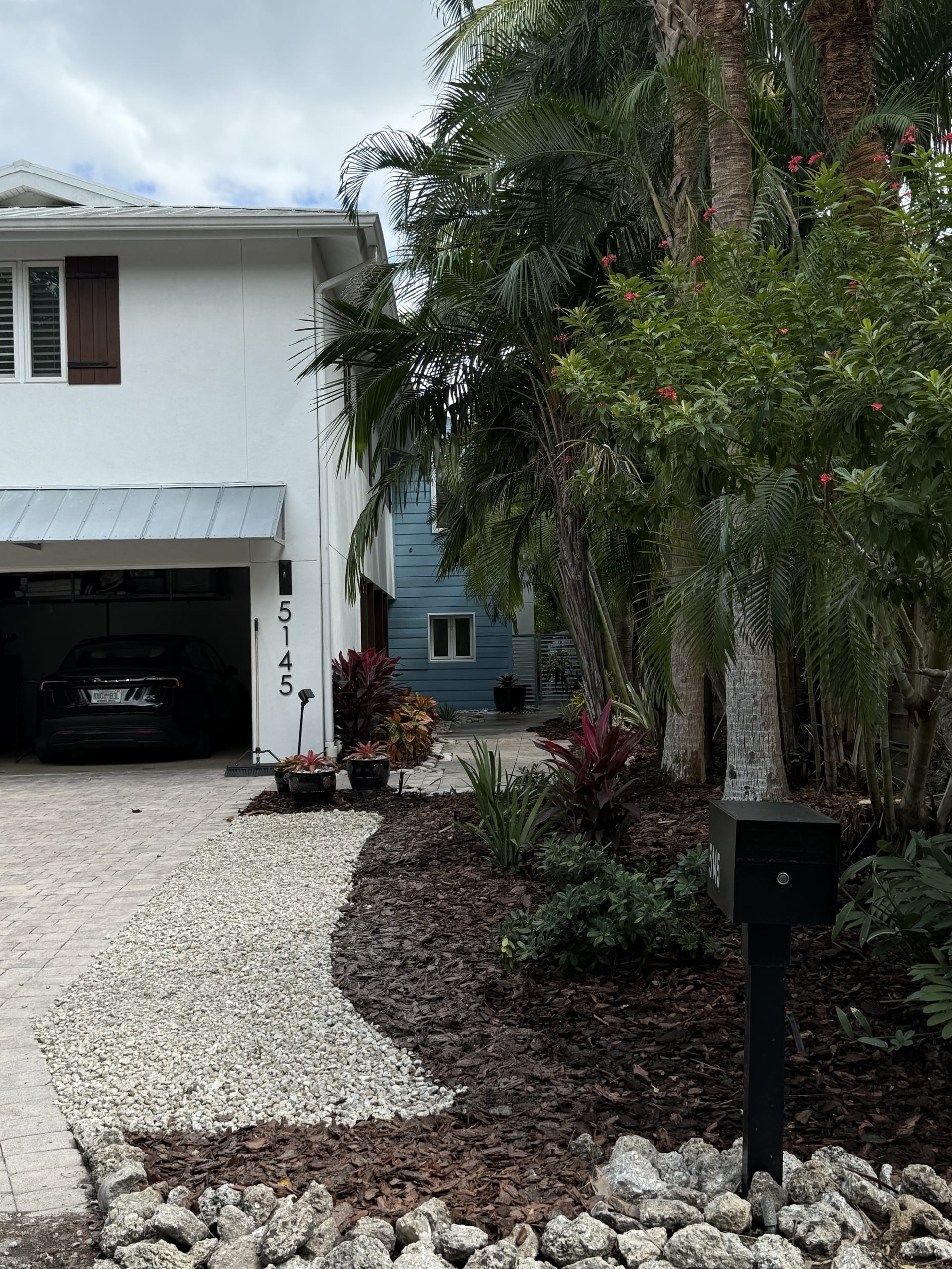 After: the same two-story home with finished tropical beds, palms, and paver driveway